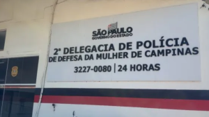 importunação sexual em Campinas: Placa de identificação da 2ª Delegacia de Polícia de Defesa da Mulher de Campinas, com telefones e indicação de atendimento 24 horas, em cenário institucional.