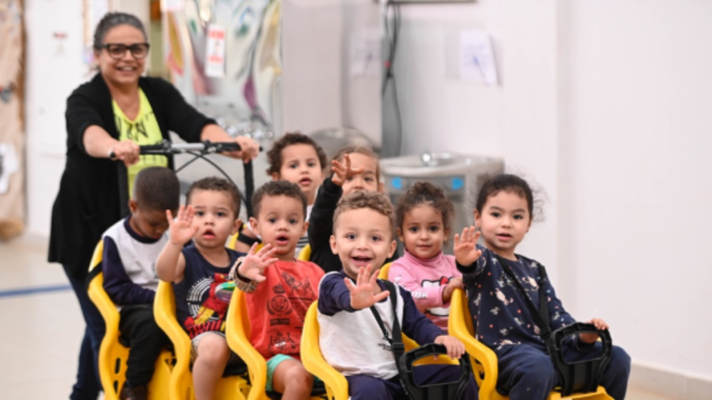 Professora acompanha alunos em sala de aula, com crianças sentadas em cadeiras amarelas e acenando para a câmera, em Campinas durante o segundo semestre letivo.