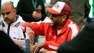 Neymar em mesa de pôquer durante competição, usando boné e jaqueta esportiva vermelha, sorrindo enquanto organiza fichas e cartas.