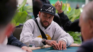 Neymar poker: jogador sentado em mesa de pôquer, usando boné preto e hoodie cinza, analisando a mão durante torneio sem premiação após eliminação.