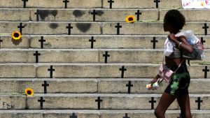 Mulher caminha em frente a degraus de cemitério com cruzes, flores e um fundo urbano, simbolizando contexto sobre mortes violentas e feminicídios.