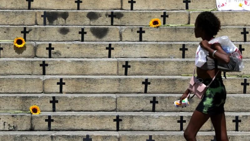 Mulher caminha em frente a degraus de cemitério com cruzes, flores e um fundo urbano, simbolizando contexto sobre mortes violentas e feminicídios.