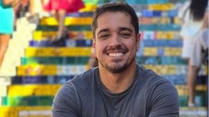 Caíque Verli sorri em foto de perfil em ambiente com fundo colorido de prateleiras e caixas, imagem usada em reportagem sobre repórter da Globo encontrado morto em apartamento