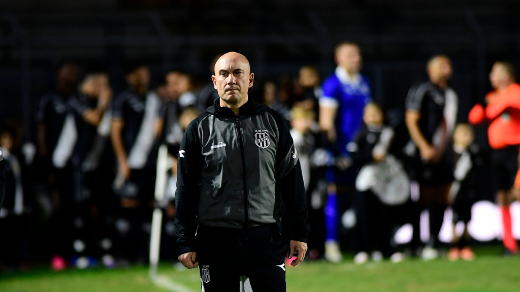 Márcio Zanardi, técnico da Ponte Preta, em campo durante partida de futebol, com jogadores desfocados ao fundo e registro de foto esportiva.