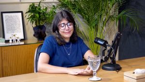 vítima de abusos: Mulher sentada em uma sala de entrevistas, com óculos e roupa azul, falando em ambiente de trabalho com microfone e taça sobre a mesa; reportagem sobre a luta na Justiça para não ter mãe.
