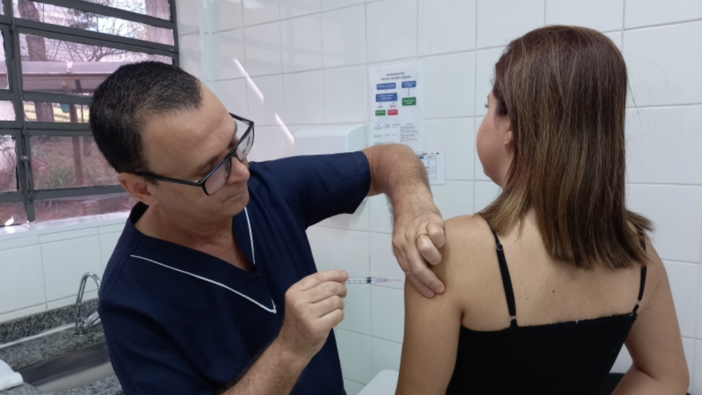 serviços gratuitos: Atendimento de saúde em programação gratuita: profissional aplica procedimento no ombro de uma mulher em unidade com parede branca e materiais de orientação ao fundo.