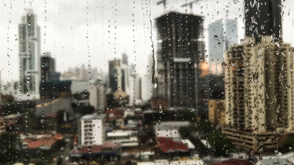 Previsão do tempo em Santos: janela com chuva e gotas no vidro sobre prédios ao fundo, indicando chuva e queda nas temperaturas neste fim de semana.