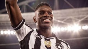 Luiz Henrique em campo, com uniforme do Botafogo, comemorando com os braços erguidos em estádio iluminado ao fundo, em destaque para notícia na Rússia.