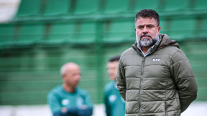 Rômulo Amaro, presidente do Guarani, aparece em foto na área técnica do clube, com expressão séria, ao lado de um banner verde ao fundo durante reeleição após impugnação de chapa da oposição.