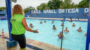 Guarujá, instruções de cursos e atividades: instrutora de camisa verde orienta turma em piscina ao ar livre, com placa “CAEE ISABEL ORTEGA” ao fundo e cenário de aula aquática.