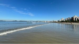 Vista da orla da Baixada Santista com mar calmo e prédios ao fundo, céu azul e previsão de calor; tempo na Baixada Santista com 32 °C antes da virada no tempo.