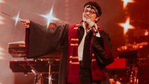 Bruno Mars surge em show usando fantasia de Harry Potter, com óculos e capa listrada vermelha e preta, cantando ao microfone sob luzes de palco