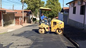 Operação Tapa-Buraco em Piracicaba: equipamento de asfalto com trabalhador realizando recuperação de via com nova massa na rua, em área residencial da cidade.