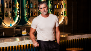 Reynaldo Gianecchini em novo visual, posando em um bar sofisticado com iluminação quente e fundo com bebidas e garrafas atrás