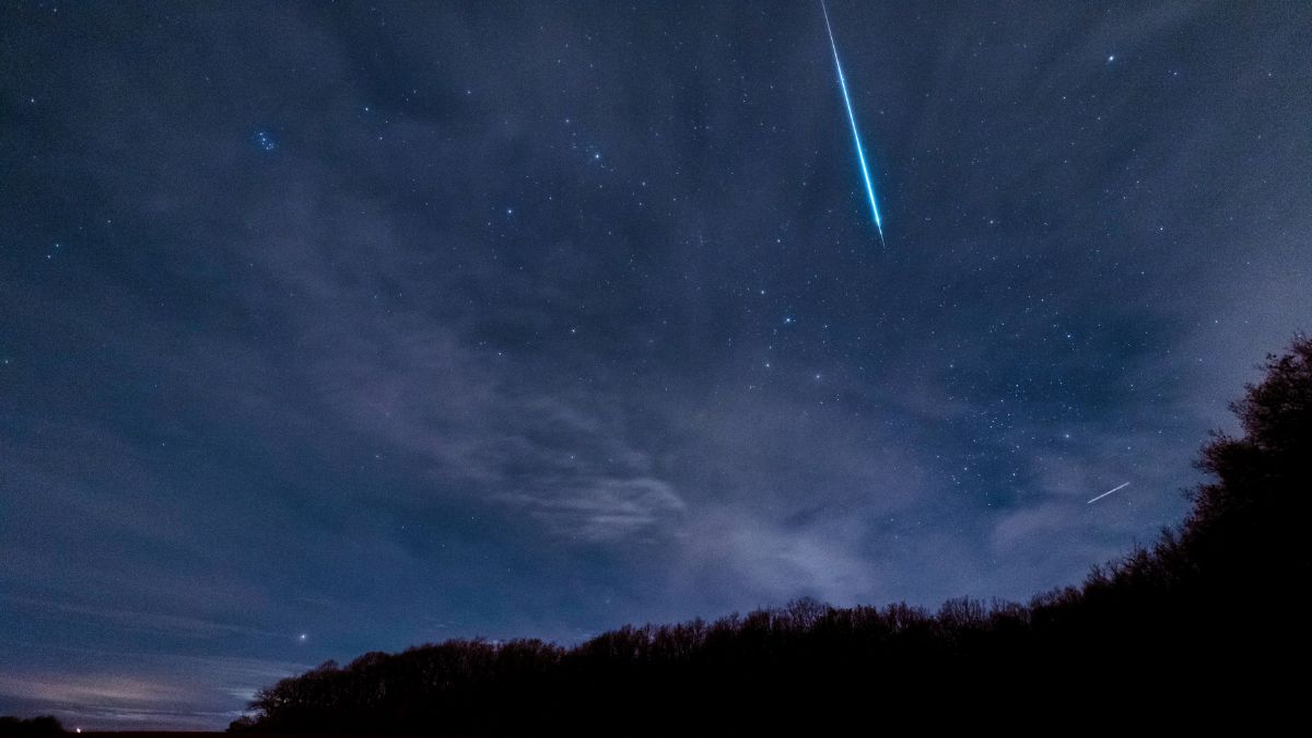Chuva de meteoros ilumina o céu noturno, com trilhas luminosas atravessando as estrelas e nuvens ao fundo, vista a partir de árvores e horizonte escuro.