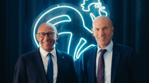 Zidane França é mostrado em foto ao lado de outra pessoa, ambos em traje social, sorrindo diante de cenário com luzes em formato de silhueta de torcedor e símbolos da França. A imagem destaca a confirmação de Zidane como novo técnico da seleção.