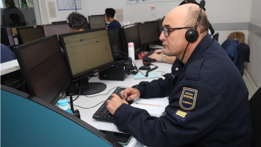 Atendente usando headset em central de atendimento, com uniforme da Guarda Civil/Guarda Municipal de Campinas, enquanto usa computadores; imagem sobre alerta de golpe em Campinas envolvendo criminosos se passando pelo órgão.