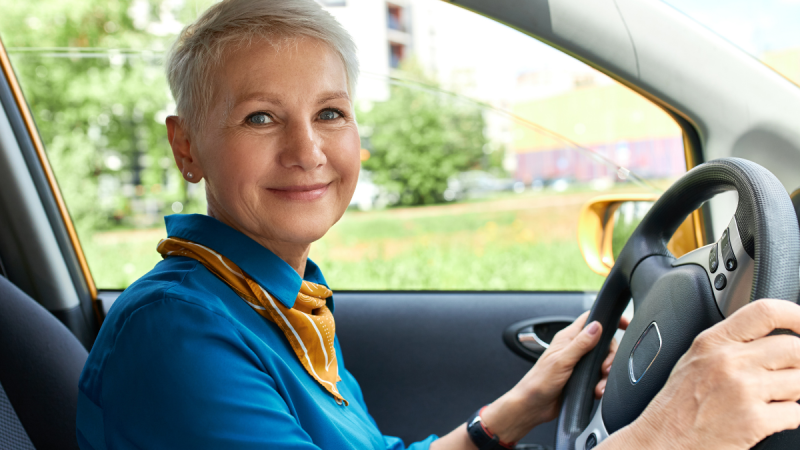 Idosa ao volante dentro do carro, pronta para dirigir, em imagem que destaca a Credencial de estacionamento para idosos e a emissão online gratuita.