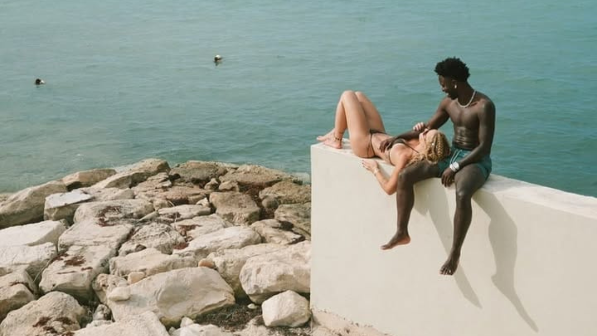 Virginia retorna de viagem com Vini Jr e faz revelação aos seguidores, com o casal descansando em uma paisagem à beira-mar, sentado em blocos de pedra e observando o oceano