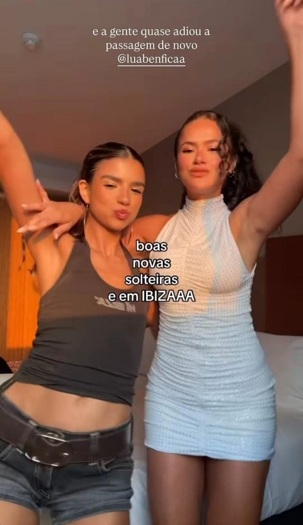 Duas mulheres posam em uma sala, uma ao lado da outra, com os braços erguidos. O texto na imagem diz “boas novas solteiras e em IBIZAAA”, além de @luabenficaa na legenda.