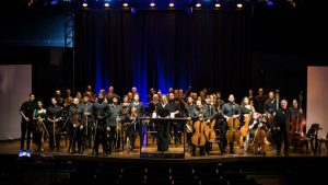 Foto de grupo de músicos em palco, com a Orquestra Filarmônica Inclusiva em Campinas durante concerto sob luzes azuis, com instrumentos de cordas e sopros.