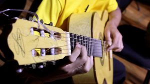 Núcleo Artístico Palácio das Artes: Mãos de músico tocando guitarra em apresentação de choro e bossa nova, com foco nas cordas e no instrumento; Praia Grande anuncia inscrições gratuitas.