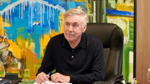 Ancelotti sentado em uma cadeira, em entrevista, com mãos apoiadas sobre uma mesa, ao fundo há um cenário colorido, projetando conversa sobre retorno ao Brasil após a Copa do Mundo.