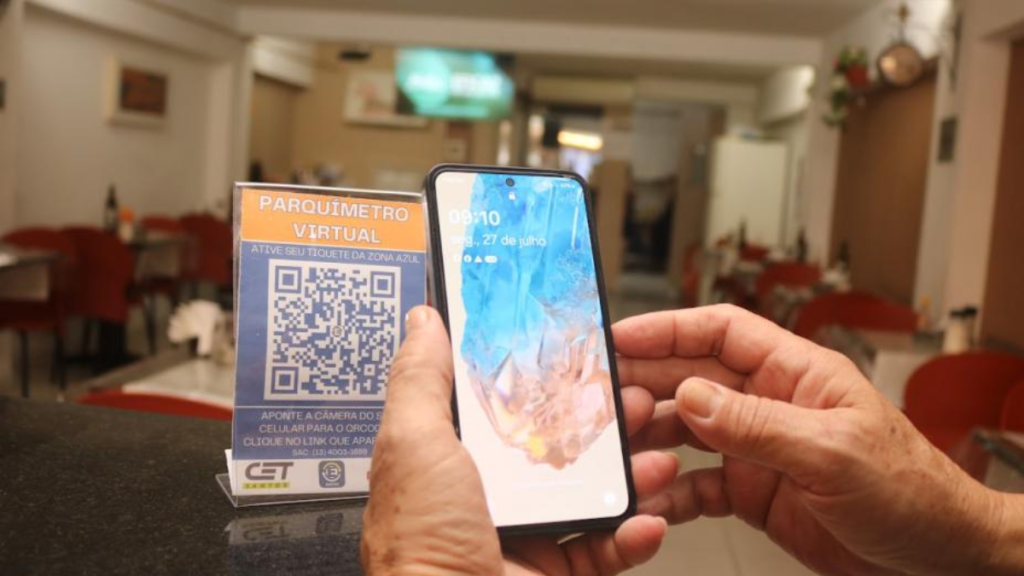 Pagamento da Zona Azul por QR Code em comércio: pessoa aponta o celular com tela de QR para realizar o estacionamento, com cartaz “PARQUIMETRO VIRTUAL”. Crédito Prefeitura de Santos