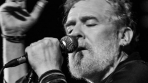 Glen Hansard ao microfone em apresentação musical em preto e branco, com olhar concentrado e mão próxima ao equipamento no palco.