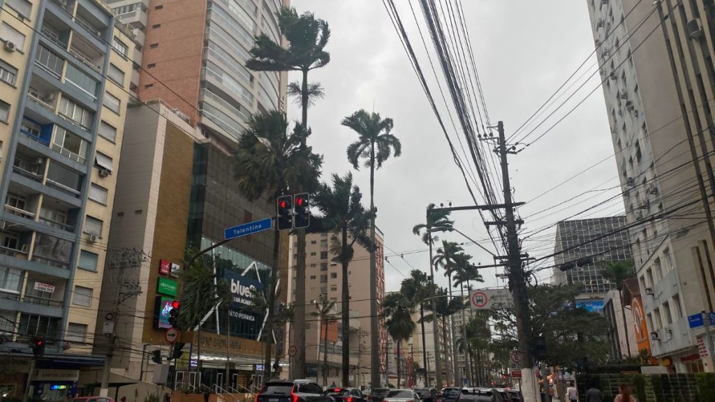 Alerta do Inmet prevê chuva intensa e ventos fortes em regiões de São Paulo; cena urbana com prédios altos, carros e palmeiras sob céu nublado e linhas de energia.