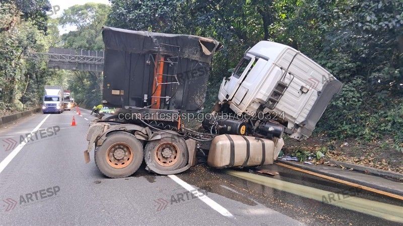 Anchieta: Registro do acidente em Cubatão: caminhão bitrem tombado na rodovia, com cabine danificada e carga ao lado da mureta de proteção, em meio à vegetação. Foto: Reprodução/ARTESP.