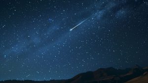 Céu noturno com estrelas e uma trilha luminosa de meteoro atravessando a Via Láctea, acima de montanhas em um horizonte escuro; chuva de meteoros poderá ser vista nesta sexta.