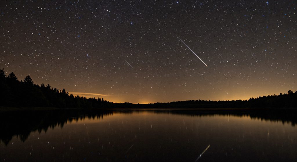 No céu escuro e estrelado acima de um lago, destacam-se rastros de meteoros e uma estrela cadente, ilustrando a chuva de meteoros