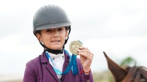 Atleta de salto usando capacete e traje equestre segura uma medalha dourada em cenário ao ar livre, destacando o Campeonato Brasileiro de Salto Iniciante 2026 com recorde de participantes.