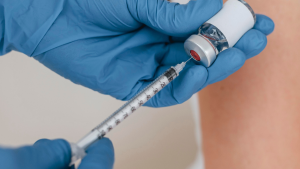 Profissional de saúde com luvas azuis aplicando uma vacina com seringa no braço, em campanha do Governo de SP para ampliar vacinação contra sarampo em 645 cidades.