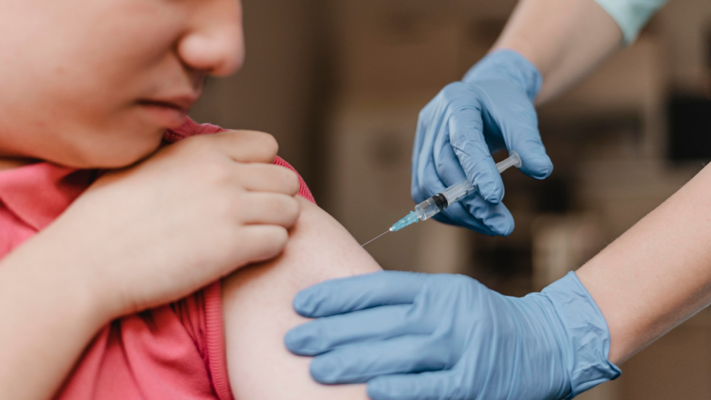 Criança sendo vacinada em posto de saúde, com profissional de luvas aplicando injeção e responsável ao lado, reforçando cuidados de vacinação contra sarampo com caderneta de vacinação