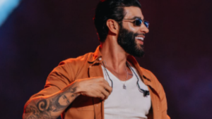 Gusttavo Lima em apresentação ao vivo, usando óculos escuros e jaqueta laranja sobre camiseta branca, com tatuagens no braço e expressão sorridente ao fundo desfocado.