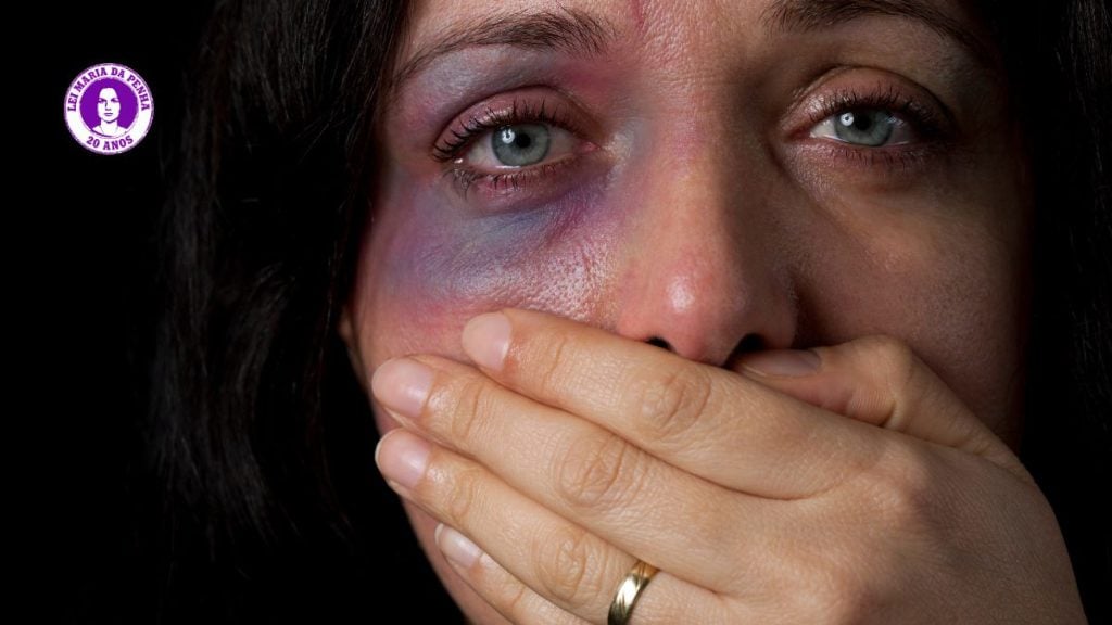 Mulher com a mão cobrindo o rosto, olhos sobre a câmera e expressão de sofrimento, em fundo escuro; recurso de conscientização sobre Lei Maria da Penha e violência contra a mulher.