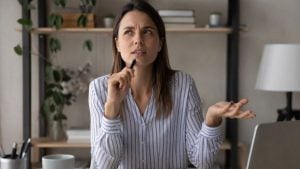 esqueci a fala: Mulher em ambiente doméstico, falando e gesticulando com a mão, com expressão de dúvida, ao pensar por que esquece a fala no meio da conversa enquanto tenta lembrar o que ia dizer