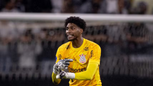 Hugo Souza, goleiro do Corinthians, aparece em campo usando uniforme amarelo, com luvas e olhar atento, durante partida no estádio; ele é citado em reportagem sobre acusações após comentário envolvendo o São Paulo.