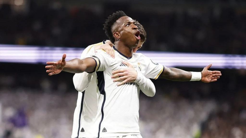 Vini Jr comemora um gol com os braços abertos usando o uniforme do Real Madrid em partida no estádio, em foto de reprodução.