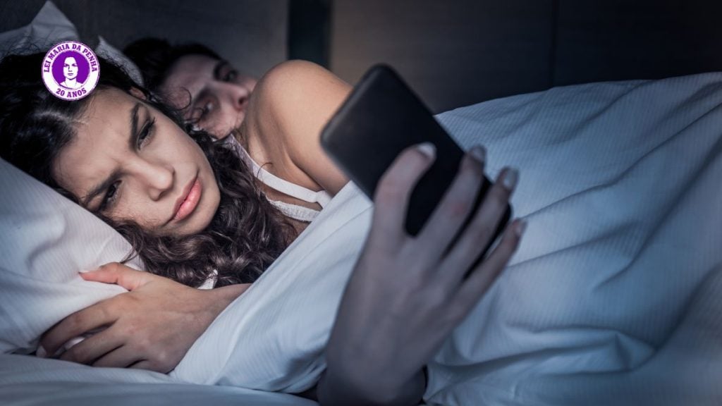 Imagem com casal na cama em clima tenso, com a mulher deitada e o homem ao fundo, sugerindo ameaça e alerta sobre violência contra a mulher e suas diferentes formas