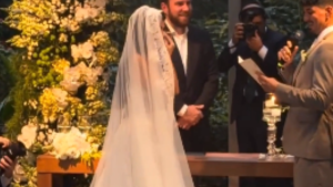 Gabriel Medina e Isabella Arantes aparecem em evento ao ar livre, com Medina vestido de branco e Isabella ao lado, enquanto fotógrafos registram a ocasião. A imagem remete à revelação de gravidez e ao nome do primeiro filho.