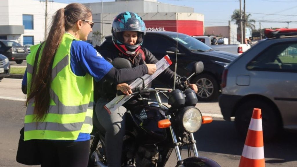 Agente de trânsito em colete refletivo orienta motociclista com capacete em uma ação educativa na via, com cones de sinalização e veículos ao fundo; no primeiro semestre de 2026 foram realizadas 166 ações educativas (Divulgação/Emdec). Inclui o tema Mortes no trânsito em Campinas.