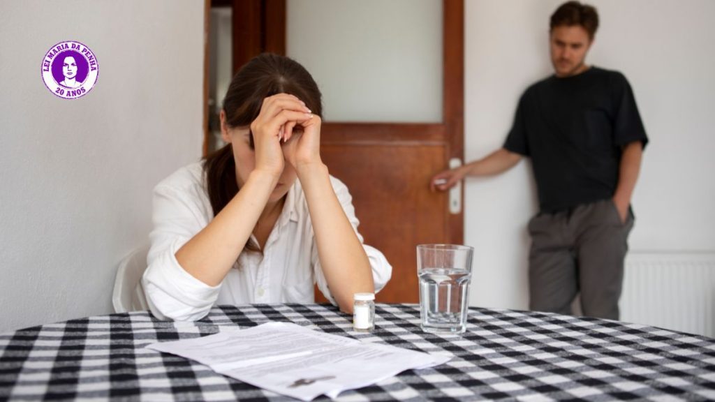 Mulher com as mãos no rosto sentada à mesa, enquanto um homem atrás da porta observa com expressão séria; imagem sobre violência contra a mulher e alerta às cinco caras da violência.