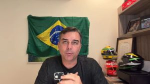 Flávio Bolsonaro vaquinha online: homem em ambiente interno fala para a câmera com bandeira do Brasil ao fundo, em contexto de lançamento de vaquinha para financiar campanha presidencial.