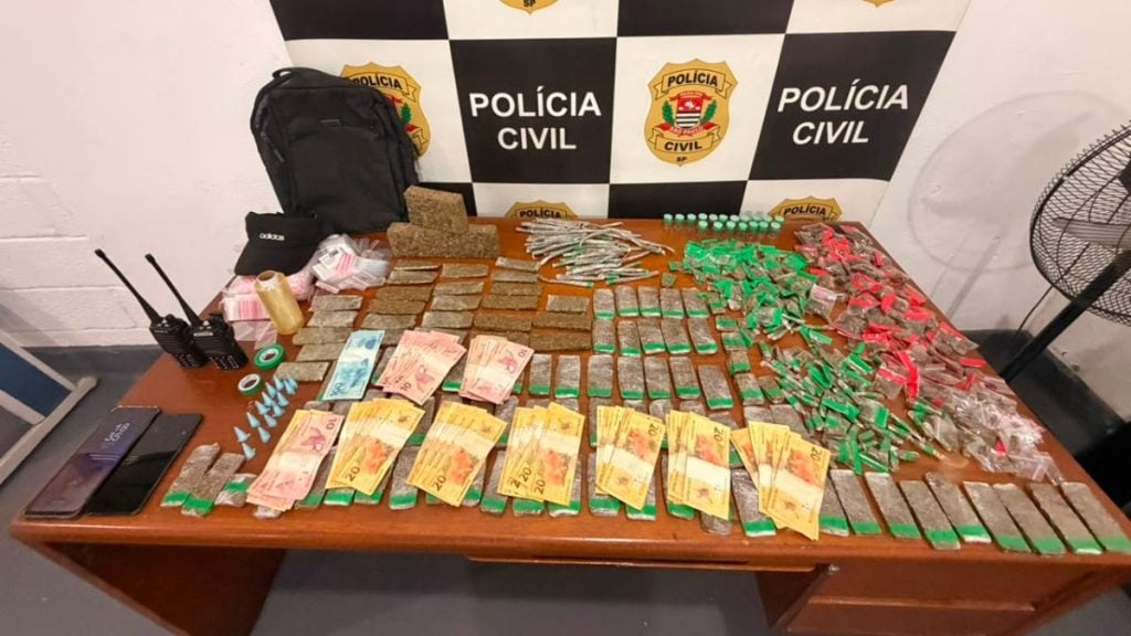 Preso roubo Santos: Foto da Polícia Civil com materiais apreendidos em operação: mesa com drogas, dinheiro em cédulas, celulares e rádios comunicadores, ao fundo a identificação Policia Civil.