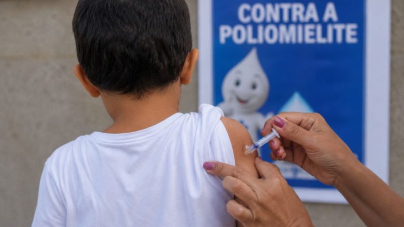 Dia D da multivacinação: criança sendo vacinada com seringa na mão e banner de campanha contra a poliomielite ao fundo, em atendimento em todo o país neste sábado (22).