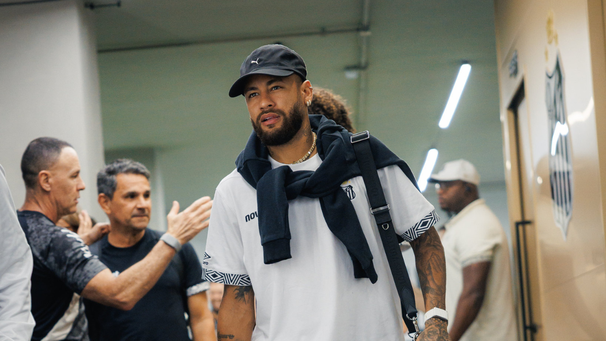 Neymar em cenário de bastidores, usando boné e moletom com mochila, enquanto pessoas ao fundo parecem conversar ou aguardar. Foto em ambiente fechado.