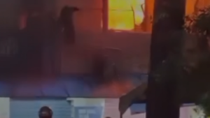 Cena noturna com iluminação vermelha e fumaça sugerindo incêndio em residência no Guarujá, com pessoas na área ao fundo e marco de janela ao centro